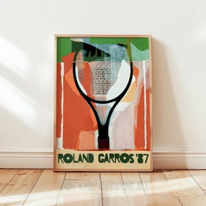 May include: Framed art print featuring a tennis racket design. The artwork has a vibrant color palette with orange, green, and white hues. The text "ROLAND GARROS '87" is displayed at the bottom in a green font. The print is displayed on a wooden floor.