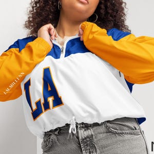 Puede incluir: Chaqueta blanca, azul y dorada con las letras "LA" en azul. La chaqueta tiene un cordón en la cintura y cierre de media cremallera. Las mangas son doradas y los hombros azules. El texto "I.M.MILLENN" está impreso en la manga izquierda.