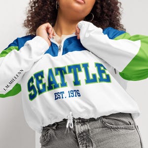 May include: A white, blue, and green color-blocked quarter-zip pullover jacket featuring the text SEATTLE EST. 1976 in blue and green lettering on the front. The jacket has long sleeves, a drawstring hem, and the text I.M.MILLENN on the sleeve.