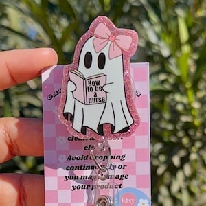 May include: A pink glitter ghost with a pink bow holding a book that says "How to be a nurse". The ghost is attached to a clear plastic badge holder with a retractable reel. The badge holder has a pink and white checkered background with a daisy pattern and the word "Etsy" printed on it.