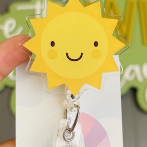May include: A yellow sun with a smiling face on a clear plastic badge holder with a retractable badge reel. The badge reel is attached to a pink and purple striped ribbon.
