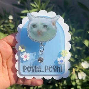 May include: A light blue retractable badge holder with a crying cat face design. The badge holder has a white scalloped border, small flower accents, and the text "Poshi_Poshi".