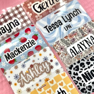 May include: A collection of name badges with various patterns and names. Designs include gingham, floral, strawberries, and leopard print. Names such as Genna, Tessa Lynch, Mackenzie, Ashley, and Elizabeth are visible. Each badge is in a clear plastic sleeve.