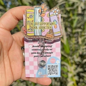 May include: A clear plastic badge holder with a gold glitter background. The holder features a stack of colorful books with the text "HERE TO SUPPORT MY BOOK ADDICTION". The holder is attached to a retractable badge reel with a clear plastic clip.
