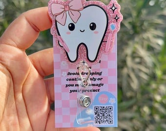 Puede incluir: Portagafete en forma de diente rosa con purpurina, con un lazo y un amuleto de cepillo de dientes. El portagafete tiene un carrete retráctil de plástico transparente y un fondo a cuadros rosa y blanco con el texto "Evita que se caiga o que se toque bruscamente, ya que podrías dañarlo." y un código QR.