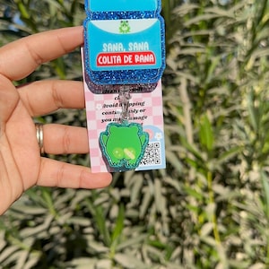 May include: A blue and green glitter charm with a frog design. The charm has a clear plastic backing and a metal ring. The charm says "Sana, Sana Colita de Rana" in Spanish.