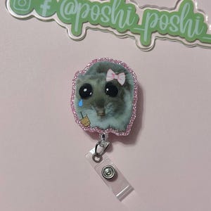 May include: A pink glitter badge holder with a cartoon image of a hamster wearing a pink bow and holding a cup of iced coffee. The hamster has large eyes and a sad expression. The badge holder has a clear plastic clip.