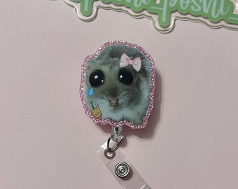 May include: A pink glitter badge holder with a cartoon image of a hamster wearing a pink bow and holding a cup of iced coffee. The hamster has large eyes and a sad expression. The badge holder has a clear plastic clip.