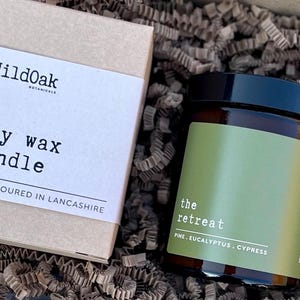 May include: A soy wax candle in an amber glass jar with a black lid and a green label reading "the retreat" and "PINE. EUCALYPTUS. CYPRESS," alongside a tan cardboard box labeled "WildOak" and "soy wax candle hand poured in Lancashire."