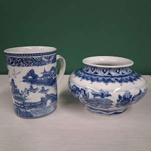 Könnte beinhalten: Eine weiße Keramiktasse und -vase mit blauen Landschaftsdesigns. Die Tasse hat einen Henkel und eine detaillierte Szene einer Brücke und von Gebäuden. Die Vase ist rund mit einer weiten Öffnung und einem ähnlichen blauen Muster.