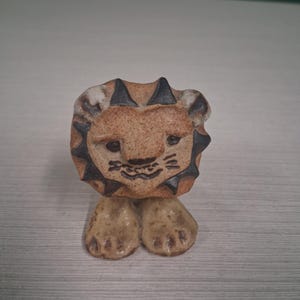 May include: A ceramic lion figurine with a tan body, a detailed face, and black accents on the mane. The lion has a friendly expression and is standing on two small feet. The figurine is a decorative item.
