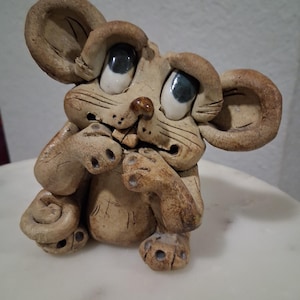 May include: A whimsical ceramic mouse figurine with large, expressive eyes and oversized ears. The mouse is light brown with darker accents, and its paws are curled. The figurine has a slightly distressed, handmade appearance, adding to its charm.
