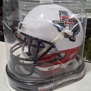 Puede incluir: Un casco de fútbol americano blanco y rojo con una máscara negra, exhibido en una caja de plástico transparente. El casco presenta un logotipo del equipo y el número 5. La marca "Schutt" es visible en la caja.