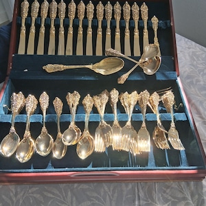 May include: A gold-toned cutlery set housed in a dark wooden storage case with a dark blue velvet interior. The set includes numerous knives, spoons, and forks, all featuring ornate, embossed floral detailing on the handles of each piece.