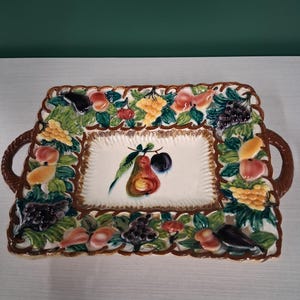 Pode incluir: Bandeja retangular de cerâmica com centro branco e borda marrom. A borda é decorada com frutas coloridas e folhas verdes. A bandeja tem duas alças marrons e um desenho de pera e ameixa pintado no centro.