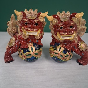 May include: Pair of red ceramic guardian lion statues. Each statue has a fierce expression, with gold accents on the mane, wings, and base. The base is a blue and gold sphere. The statues are on a white surface.