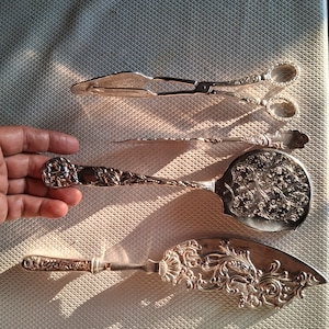 Puede incluir: Un surtido de utensilios de servir de plata antiguos, que incluyen pinzas, una cuchara de servir delgada y un servidor de pasteles. Las piezas presentan diseños florales y de volutas ornamentados, lo que refleja una estética vintage. Los artículos se exhiben sobre una superficie texturizada.