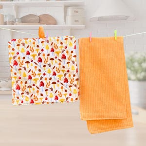 May include: Two kitchen towels hanging on a clothesline. One is patterned with red, yellow, and brown mushrooms and leaves on a white background. The other is a solid orange corduroy towel.