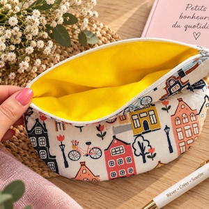 Pode incluir: Uma bolsa de tecido semicircular com fecho de correr branco e forro interior amarelo brilhante. O exterior apresenta um padrão repetitivo de casas coloridas, bicicletas e flores sobre um fundo branco. A bolsa é mantida aberta.
