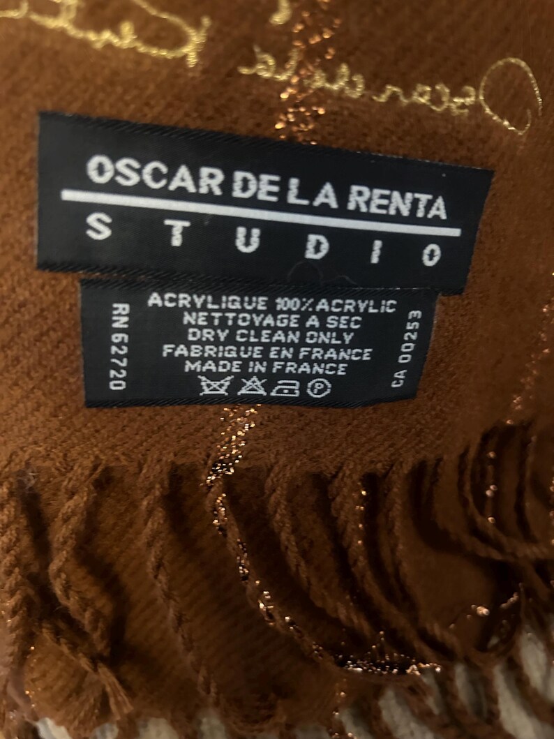 Pode incluir: Cachecol castanho e dourado com franjas. O r&oacute;tulo diz "Oscar de la Renta Studio" e inclui instru&ccedil;&otilde;es de cuidados em franc&ecirc;s e ingl&ecirc;s.