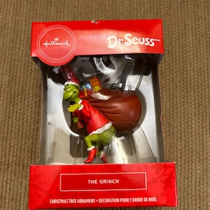 May include: A Hallmark Christmas tree ornament featuring the Grinch from Dr. Seuss. The Grinch is green with a red Santa suit and hat, carrying a brown sack. The ornament is in a red box with the text "THE GRINCH".