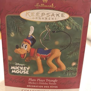 May include: A Hallmark Keepsake ornament featuring Pluto from Disney's Mickey Mouse. Pluto is depicted playing a triangle, wearing a blue and red uniform. The ornament is part of the Mickey's Holiday Parade Collector's Series.
