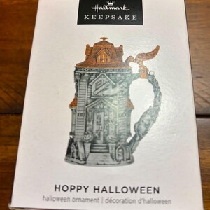 May include: A white Hallmark Keepsake box featuring a graphic of a gray haunted house beer stein ornament with orange roof accents and a handle shaped like a raven. The text reads Hallmark KEEPSAKE, HOPPY HALLOWEEN, and halloween ornament.