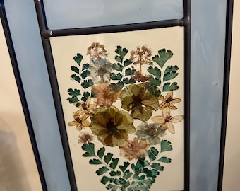 Vitral com flores secas penduradas na parede. As flores são tons de rosa claro e cores terrosas com hastes verdes. Borda de vitral azul.