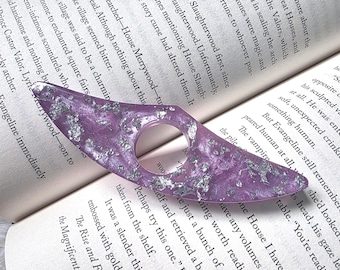 May include: A purple resin book page holder with silver foil accents. The reading accessory has a rounded hole for the finger and a wing-like shape. The book is open, displaying text.