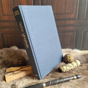 May include: A blue hardcover book titled "Sefer Yetzirah" by Aryeh Kaplan. The book is propped up on a fur-like surface, with a wooden wand, sage bundle, and other items nearby. The book's title and author are in gold lettering.