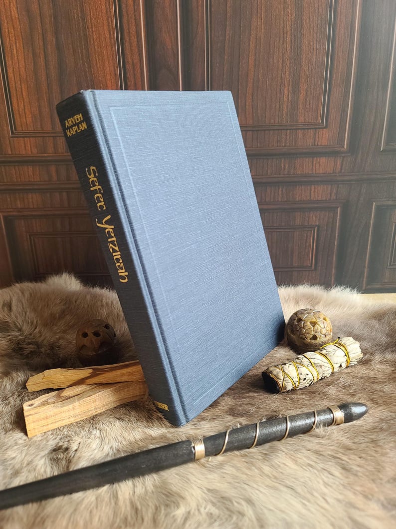 May include: A blue hardcover book titled "Sefer Yetzirah" by Aryeh Kaplan. The book is propped up on a fur-like surface, with a wooden wand, sage bundle, and other items nearby. The book's title and author are in gold lettering.