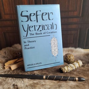 May include: A light blue book titled "Sefer Yetzicah, The Book of Creation" with the author Aryeh Kaplan. A black wand with copper wire, a sage bundle, and a carved stone sphere rest on a fur surface.