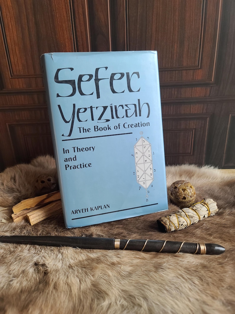 May include: A light blue book titled "Sefer Yetzicah, The Book of Creation" with the author Aryeh Kaplan. A black wand with copper wire, a sage bundle, and a carved stone sphere rest on a fur surface.