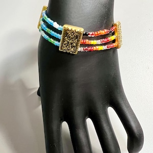 Pode incluir: Pulseira de contas multicoloridas com detalhes dourados. A pulseira apresenta quatro filas de contas em tons de azul, verde, vermelho, laranja, amarelo e branco. Os detalhes dourados são retangulares e têm um design decorativo.