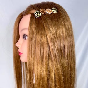 Könnte beinhalten: Eine Haarspange mit vier kreisförmigen Dekorationselementen. Die Elemente weisen Tiermuster in Schwarz, Braun und Beige auf. Die Spange ist an einem Schaufensterkopf mit langem, glattem, hellbraunem Haar befestigt.