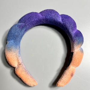 May include: A headband with a gradient of peach, blue, and purple colors. The headband is made of a soft, plush material and has a slightly ruffled texture.