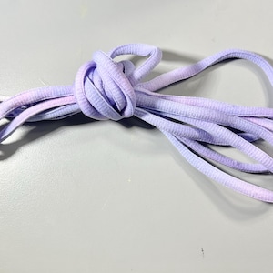 Puede incluir: Un par de cordones de zapatos de color degradado púrpura y blanco. Los cordones están atados en un nudo y listos para ser usados.
