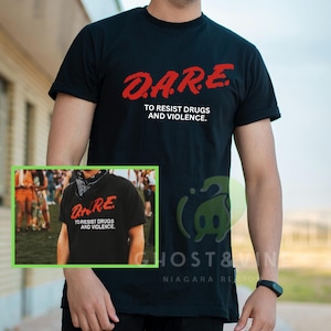May include: Black t-shirt with the red text "D.A.R.E. TO RESIST DRUGS AND VIOLENCE." The image shows a person wearing the shirt. A smaller image of the same shirt is also visible.