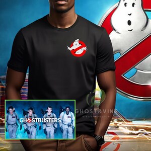 May include: A black t-shirt with a white ghostbusters logo and a red circle with a diagonal line through it. The shirt is being worn by a person. The image also shows a group of four people in ghostbusters costumes walking in front of a blue background. The text "Ghostbusters" is visible in the image.