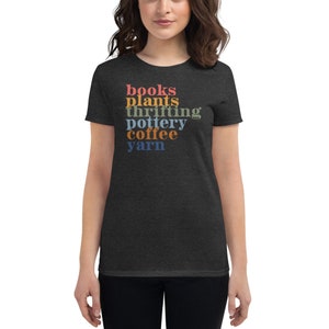 May include: Dark gray t-shirt with the words "books, plants, thrifting, pottery, coffee, yarn" in a vertical stack of colorful text. The shirt is a crew neck style and is made of a soft material.