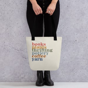 May include: Cream-colored tote bag with black handles. The bag features the words "books, plants, thrifting, pottery, coffee, yarn" in a stacked, retro-style font. The bag is held by a person wearing black pants and boots.