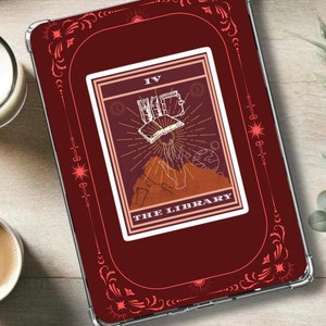 Puede incluir: Una funda de tableta burdeos con un diseño de carta del tarot que presenta un libro abierto, rayos de sol y el texto "THE LIBRARY". La funda tiene detalles florales decorativos y una cubierta protectora transparente.