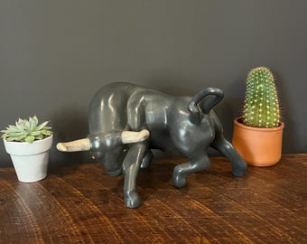 May include: A dark gray bull figurine with light-colored horns, posed in a charging stance. The sculpture is on a wooden surface, with small potted plants on either side. The background is a dark gray wall.