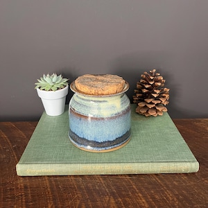 May include: A handcrafted ceramic jar with a cork lid. The jar features a blue and brown glaze, with a light yellow band near the top. It sits on a green book, next to a small succulent in a white pot and a pinecone. Home decor.
