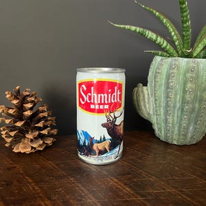 May include: A vintage Schmidt Beer can with a red and white label. The label features the brand name and an illustration of elk in a mountain landscape. The can is silver with a red top and is displayed on a wooden surface.