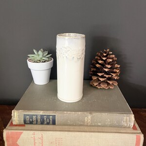 May include: A white ceramic vase with a textured, lace-like design around the top. The vase is cylindrical and sits on a stack of vintage books. A small potted succulent and a pinecone are also present.