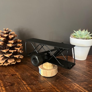 May include: A black metal airplane sculpture, mounted on a wooden base. The biplane design features a detailed frame and a nut as the propeller. The sculpture is displayed on a wooden surface, with a pinecone and a succulent in the background.