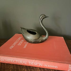 May include: A black cast iron swan figurine sitting on top of a red book titled "Architectural Design" by Ernest Pickering. The book is on a wooden surface.