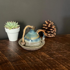 May include: A ceramic oil lamp with a rounded body, featuring a blue and brown glazed finish, a curved handle, and a thick jute wick. The lamp sits inside a matching shallow ceramic saucer, creating a cohesive set with a rustic, textured appearance.