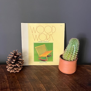 May include: A hardcover book titled "Wood Work: A Basic Manual" with an illustration of a wooden rocking chair. A pine cone and a potted cactus are on a wooden surface.